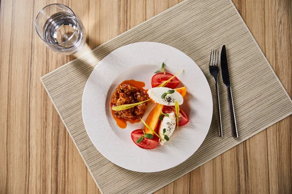 Top view of delicious restaurant dish with eggplant caviar and tomatoes served on wooden table with water and cutlery — Stock Photo