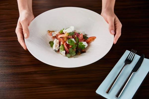 Vista recortada de la mujer sirve deliciosa ensalada de restaurante con queso en la mesa de madera con cubiertos - foto de stock