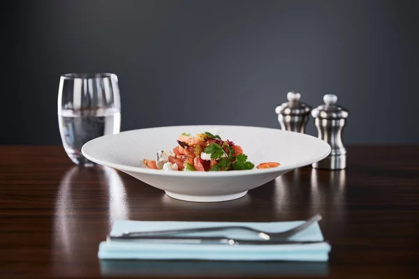 Enfoque selectivo de deliciosa ensalada de restaurante con queso servido en plato blanco en mesa de madera con agua, cubiertos y salero y pimentero sobre fondo negro - foto de stock