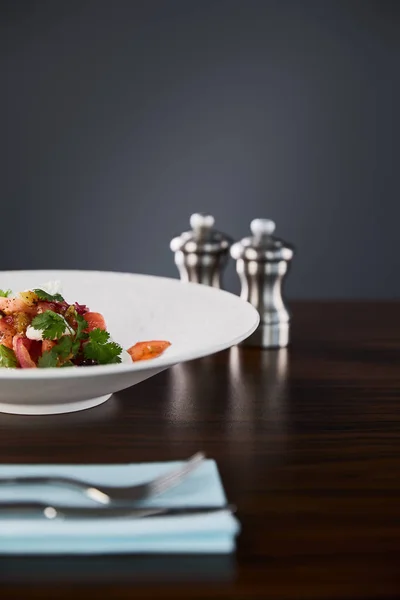 Selective focus of delicious restaurant salad with cheese served in white plate on wooden table with cutlery and salt and pepper shakers on black background — Stock Photo