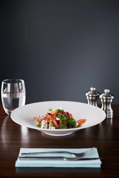 Enfoque selectivo de deliciosa ensalada de restaurante con queso servido en plato blanco en mesa de madera con agua, cubiertos y salero y pimentero sobre fondo negro - foto de stock