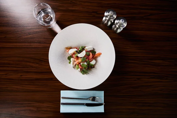 Vista superior de la deliciosa ensalada de restaurante con queso servido en plato blanco sobre mesa de madera con agua, cubiertos y salero y pimentero - foto de stock