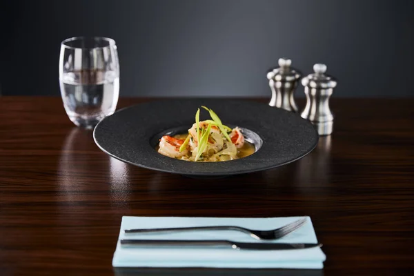 Selective focus of delicious restaurant soup with shrimps in black plate served on napkin with cutlery and water on black background — Stock Photo