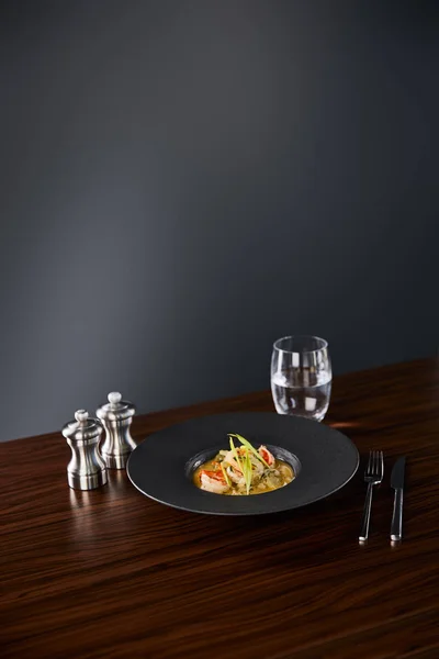 Delicious restaurant soup with shrimps in black plate served on napkin with cutlery and water on black background — Stock Photo