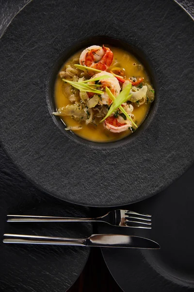 Top view of delicious restaurant soup with shrimps in black plate with cutlery — Stock Photo