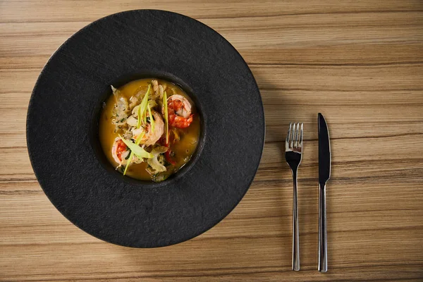 Вид на вкусный ресторанный суп с креветками в черной тарелке на деревянном столе со столовыми приборами — стоковое фото