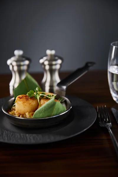 Selective focus of delicious grilled scallops with green leaves and microgreens near cutlery, water and pepper, salt shakers — Stock Photo