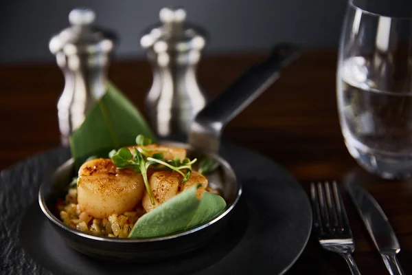 Foyer sélectif de pétoncles grillés délicieux avec des feuilles vertes et microgreens près de couverts, eau et poivre, salières — Photo de stock