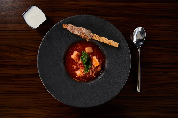 Top view of traditional ukrainian borscht garnished with parsley in black plate served with sour cream near spoon at wooden table — Stock Photo