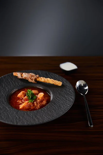 Borscht tradicional ucraniano decorado com salsa em placa preta servida com creme azedo perto da colher na mesa de madeira — Fotografia de Stock