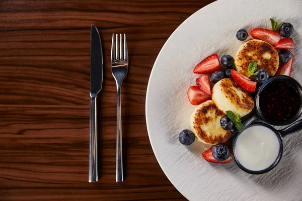 Vue de dessus de sironiki plat ukrainien traditionnel avec fraise, myrtille servie avec confiture et crème sure sur table en bois près des couverts — Photo de stock