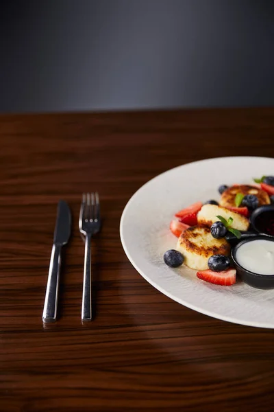 Sirniki plato tradicional ucraniano con fresa, arándano servido con mermelada y crema agria en la mesa de madera cerca de cubiertos - foto de stock