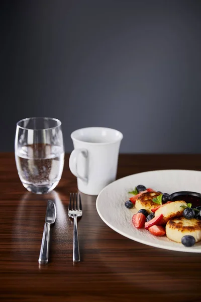 Traditional Ukrainian dish syrniki with strawberry, blueberry served with jam and sour cream on wooden table near cutlery, coffee and water — Stock Photo