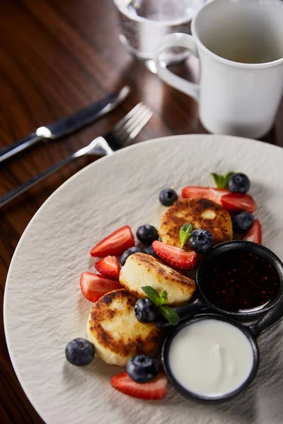 Sirniki plato tradicional ucraniano con fresa, arándano servido con mermelada y crema agria en la mesa de madera cerca de cubiertos, café y agua - foto de stock