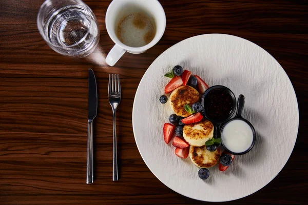 Vista superior de sirniki plato tradicional ucraniano con fresa, arándano servido con mermelada y crema agria en la mesa de madera cerca de cubiertos, café y agua - foto de stock