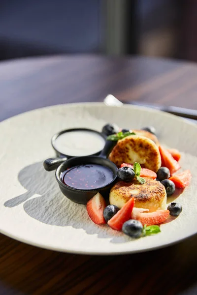 Piatto tradizionale ucraino syrniki con fragola, mirtillo servito con marmellata, panna acida sul tavolo di legno alla luce del sole — Foto stock