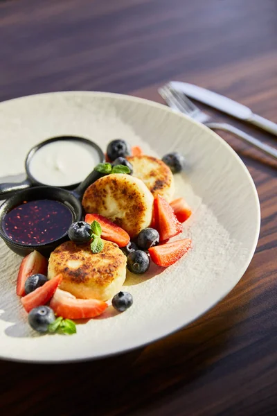 Traditional Ukrainian dish syrniki with strawberry, blueberry served with jam, sour cream and cutlery on wooden table in sunlight — Stock Photo