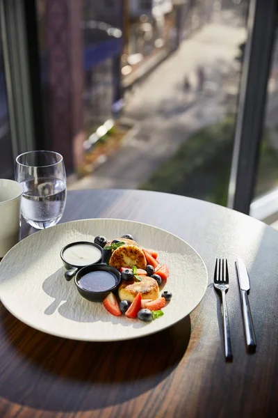 Piatto tradizionale ucraino syrniki con fragola, mirtillo servito con marmellata, panna acida, caffè, acqua e posate sul tavolo di legno alla luce del sole — Foto stock