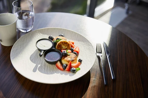 Sirniki plato tradicional ucraniano con fresa, arándano servido con mermelada, crema agria, café, agua y cubiertos en mesa de madera a la luz del sol - foto de stock