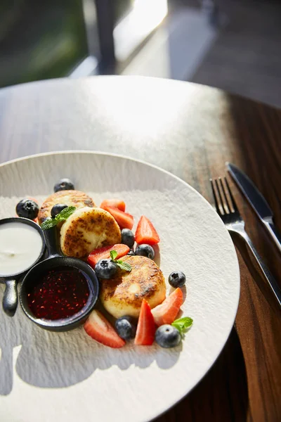 Traditional Ukrainian dish syrniki with strawberry, blueberry served with jam, sour cream and cutlery on wooden table in sunlight — Stock Photo