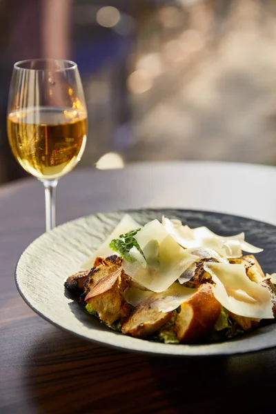 Вкусный салат Цезарь с ломтиками пармезана подается на тарелке с белым вином в солнечном свете — стоковое фото