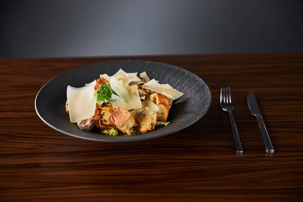 Вкусный салат Цезарь с ломтиками пармезана подается на тарелке со столовыми приборами на деревянном столе — стоковое фото