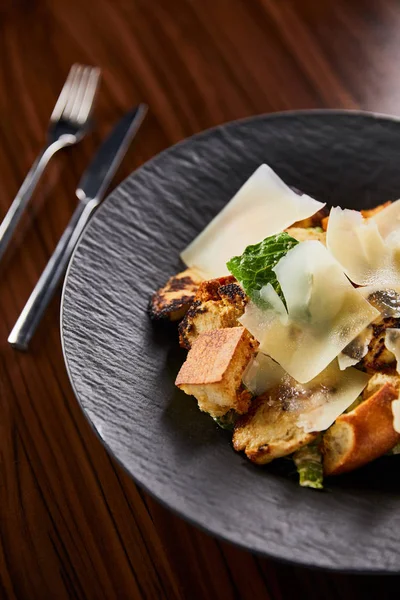 Selective focus of tasty Caesar salad with Parmesan slices served on plate with cutlery on wooden table — Stock Photo