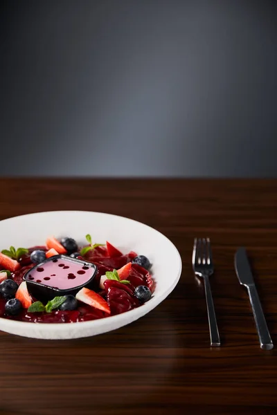 Selektiver Schwerpunkt der traditionellen ukrainischen varenyky mit Beeren in weißem Teller mit Sauce in der Nähe von Besteck auf Holztisch serviert — Stockfoto