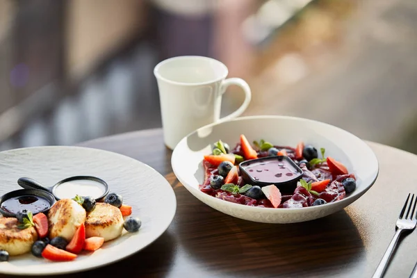 Traditional Ukrainian varenyky and syrniki with berries served in white plate with sauce near cutlery and cup on wooden table in restaurant — Stock Photo