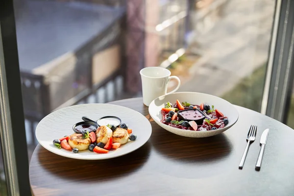 Традиційні українські вареники і сирники з ягодами подаються на білій тарілці з соусом біля столового посуду і чашкою на дерев'яному столі в ресторані. — стокове фото
