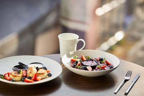 Varenyky tradicional ucraniano y syrniki con bayas servidas en plato blanco con salsa cerca de cubiertos y taza en la mesa de madera en el restaurante — Stock Photo