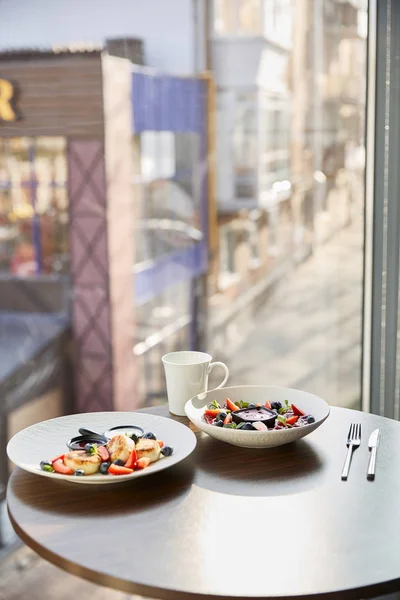 Traditional Ukrainian varenyky and syrniki with berries served in white plate with sauce near cutlery and cup on wooden table in restaurant — Stock Photo