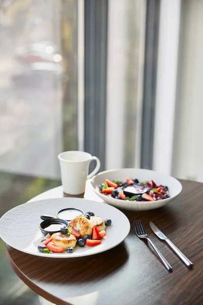 Traditional Ukrainian varenyky and syrniki with berries served in white plate with sauce near cutlery and cup on wooden table in restaurant — Stock Photo