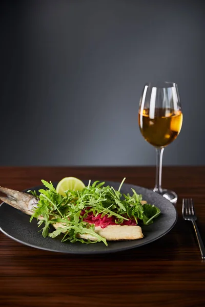 Sabroso restaurante filete de pescado con lima y rúcula en la mesa de madera cerca de tenedor y vino blanco sobre fondo negro - foto de stock