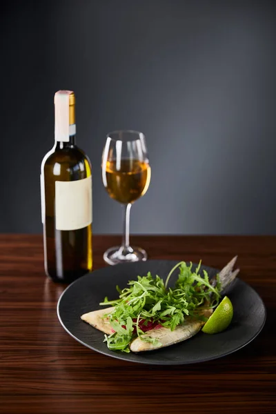 Tasty restaurant fish steak with lime and arugula on wooden table near white wine on black background — Stock Photo