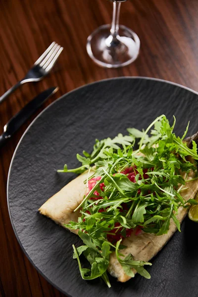 Selective focus of tasty restaurant fish steak with lime and arugula on wooden table near cutlery — Stock Photo