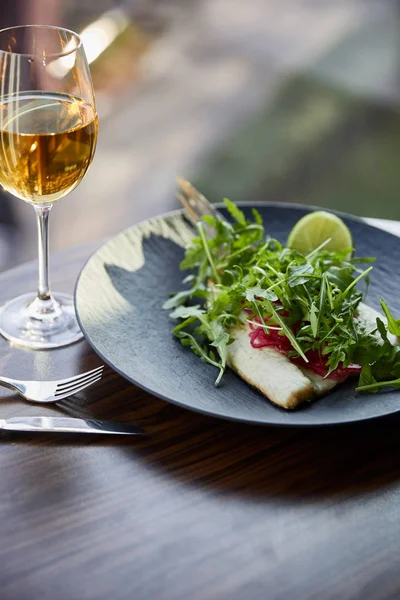Sabroso restaurante filete de pescado con lima y rúcula en la mesa de madera cerca de cubiertos y vino blanco a la luz del sol - foto de stock