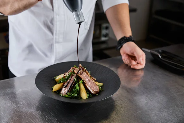 Vue partielle du chef versant la sauce sur le plat avec roquette, viande et pomme de terre à la cuisine dans le restaurant — Photo de stock