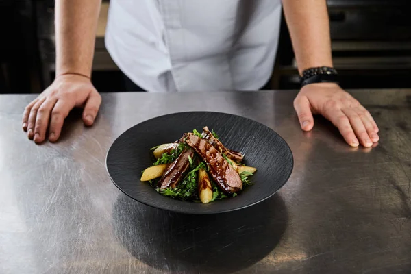 Vista parcial del chef de pie cerca del plato con rúcula, carne y patata en la cocina en el restaurante - foto de stock