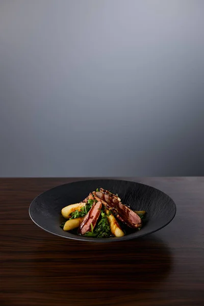 Delicious warm salad with arugula, potato and meat in black plate on wooden table on grey background — Stock Photo