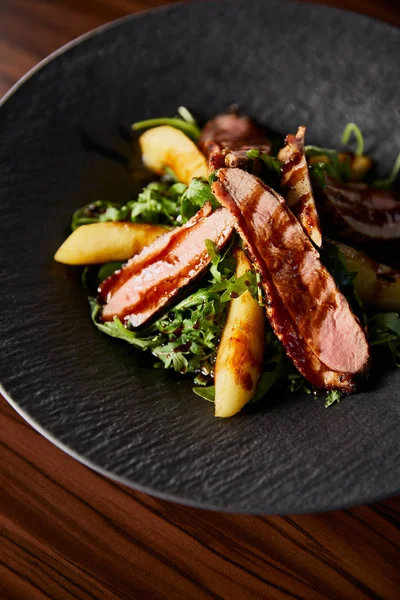 Delicious warm salad with arugula, potato and meat in black plate on wooden table — Stock Photo