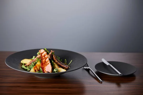 Köstlicher warmer Salat mit Rucola, Kartoffeln und Fleisch auf schwarzem Teller auf Holztisch neben Besteck auf grauem Hintergrund — Stockfoto