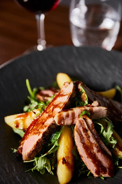 Закрытый вид на вкусный теплый салат с рукколой, картофелем и мясом в черной тарелке на деревянном столе — стоковое фото