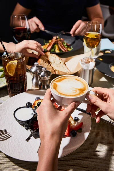 Vista recortada de la mujer comiendo delicioso syrniki y beber capuchino durante la cena con amigos en el restaurante - foto de stock