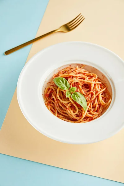 Espaguete delicioso com molho de tomate e manjericão na placa perto de garfo no fundo azul e amarelo — Fotografia de Stock