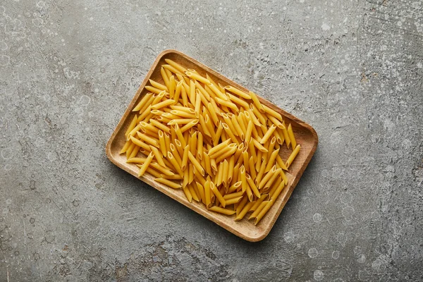 Top view of raw penne on wooden board on grey textured surface — Stock Photo