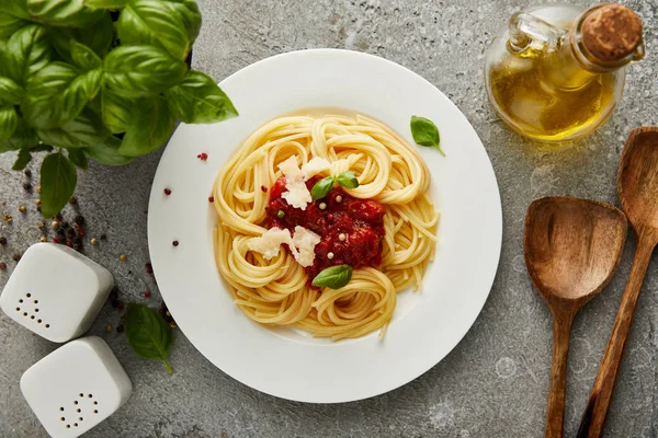 Vista dall'alto di deliziosi spaghetti con salsa di pomodoro su piatto vicino alle foglie di basilico e olio su superficie grigia testurizzata — Foto stock