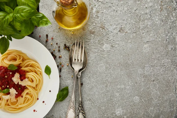 Top view of delicious spaghetti with tomato sauce on plate near basil leaves, oil and cutlery on grey textured surface with copy space — Stock Photo