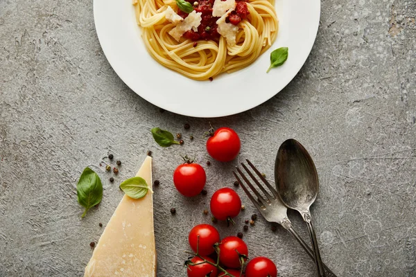 Vista superior de deliciosos espaguetis con salsa de tomate en el plato cerca de queso, tomates y cubiertos en la superficie de textura gris - foto de stock