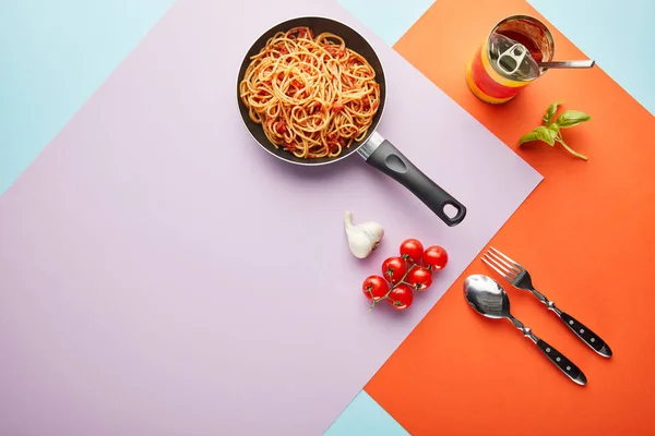 Flat lay with delicious spaghetti with tomato sauce in frying pan near ingredients on red, blue and violet background — Stock Photo